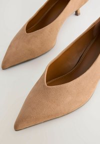 Zapatos de tacón alto de ante color beige con punta afilada y un diseño recortado. Los tacones son delgados y tienen aproximadamente 5 centímetros de altura.