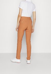 Slim-fit kalhoty v rezavé barvě s hladkou texturou, s zadními kapsami a logem, v kombinaci s bílým polo tričkem s krátkým rukávem.