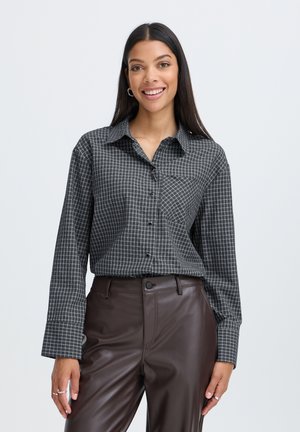 Femme aux longs cheveux noirs portant une chemise à carreaux noire et grise et un pantalon en cuir marron foncé, souriant devant un fond uni.