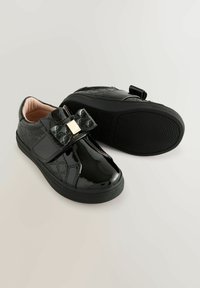 Sneakers in pelle verniciata nera con design trapuntato, dotati di un fiocco decorativo e dettagli metallici con logo. Chiusura con strap in velcro.