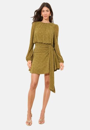 Frau trägt ein olivgrünes, strukturiertes langärmeliges Minikleid mit geraffter Taille und drapiertem Bindesdetail, kombiniert mit beigen Riemchenabsätzen.