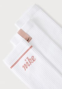 Trois chaussettes blanches Nike avec une texture côtelée, arborant un logo et une bande rose. Les chaussettes sont bien disposées.