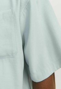 Camisa de manga corta azul claro con un bolsillo en el pecho izquierdo, que presenta una textura suave y sutiles detalles de costura.