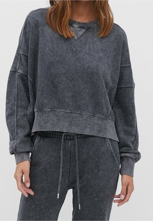 Frau trägt einen ausgewaschenen schwarzen Oversize-Sweatshirt und passende Sweatpants mit Kordelzug vor neutralem Hintergrund.