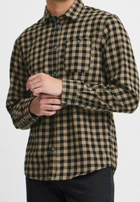 Flanelová košile s hnědočerným károvaným vzorem, zapínáním na knoflíky, jednou náprsní kapsou a kontrastními modrými knoflíky.