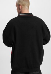 Black sweatshirt with a loose fit, features a contrast collar with red and white stripes, and a red accent at the lower side.