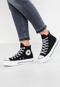 Czarne sneakersy typu high-top wykonane z płótna, z białymi gumowymi podeszwami, białymi sznurowadłami i kontrastującymi czarnymi paskami. Na boku znajduje się logo Converse.
