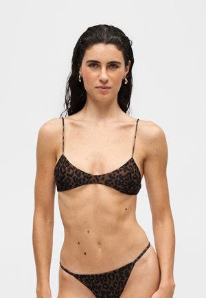 Žena s dlhými tmavými vlasmi, ktorá má na sebe hnedé a čierne bikiny s leopardím vzorom na jednofarebnom svetlom pozadí.