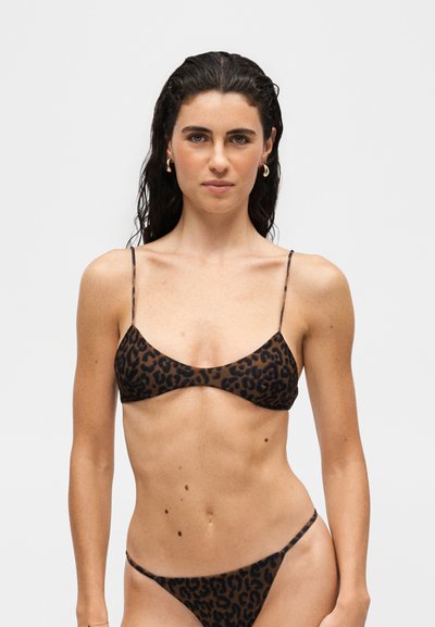 Femme aux longs cheveux foncés portant un bikini marron et noir à motif léopard sur un fond clair uni.