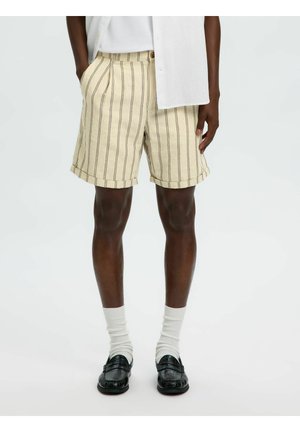 Mann trägt beige gestreifte Shorts, weiße Socken, schwarze Loafer und ein weißes Hemd mit den Händen in den Taschen vor einem schlichten Hintergrund.