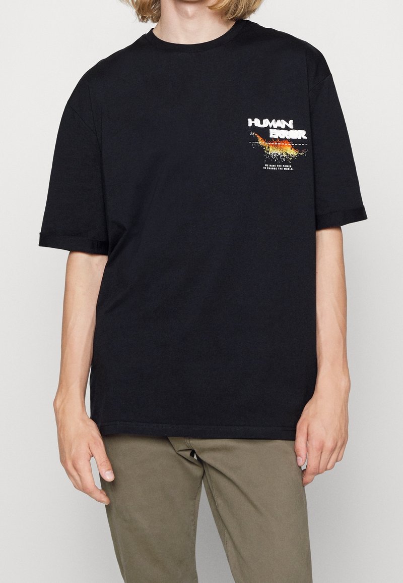 Czarny bawełniany t-shirt o luźnym kroju, z wyraźnym białym i pomarańczowym graficznym napisem po lewej stronie klatki piersiowej i krótkimi rękawami.