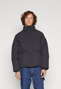 Schwarze Daunenjacke mit hohem Kragen, im boxy Schnitt, mit gestepptem Textur und minimalistischem Design. Getragen mit blauen Jeans.