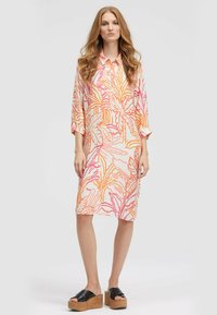 Frau mit langen, gewellten Haaren, die ein knielanges Hemdkleid mit einem orange- und pinkfarbenen tropischen Blattmuster und schwarzen Plateausandalen trägt, steht.