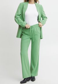 Kobieta poprawiająca talię luźnych zielonych spodni, nosząca dopasowaną zieloną marynarkę, białą koszulę i czarne szpilki, na jednolitym tle.