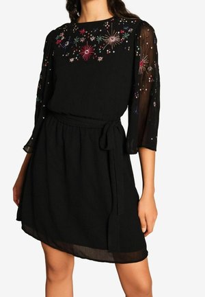 Robe noire longueur genou avec manches longues transparentes, broderie florale colorée en perles sur la poitrine et les manches, et ceinture à nouer à la taille.