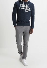 Hombre vestido con una sudadera azul marino con cordones blancos, pantalones grises con puños y zapatillas blancas, de pie contra un fondo claro y liso.