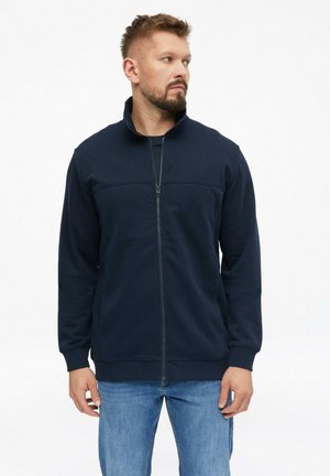 Mann mit kurzen Haaren und Bart, der eine marineblaue Jacke mit Reißverschluss und blaue Jeans trägt, steht und blickt nach links vor einem weißen Hintergrund.