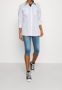 Camicia a righe con bottoni e due tasche sul petto, abbinata a pantaloni shorts in denim fino al ginocchio e sneaker nere alte.