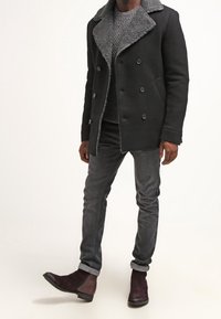 Svart ullkappa med en grå shearlingkrage, dubbelknäppt design, kombinerad med gråjeans och bordeauxfärgade ankelstövlar mot en ljus bakgrund.