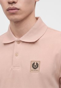 Gros plan d'une personne portant un polo rose clair à deux boutons avec un écusson du logo Belstaff sur le côté gauche de la poitrine.