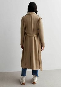 Béžový trenčkot s páskem v pase, ramenními epolety a detailem rozparku na zádech, stylizovaný na modrých džínách a bílých teniskách.