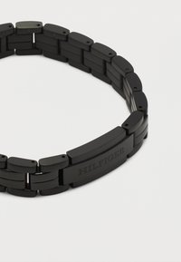 Βραχιόλι από μαύρο ματ μέταλλο με συνδετήρες και ορθογώνιο κούμπωμα χαραγμένο με τη λέξη «HILFIGER» σε λευκό φόντο.