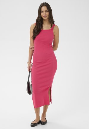 Kobieta o długich ciemnych włosach, ubrana w różową sukienkę midi bez rękawów z bocznym rozcięciem, trzymająca czarną torebkę i nosząca czarne baleriny, na białym tle.