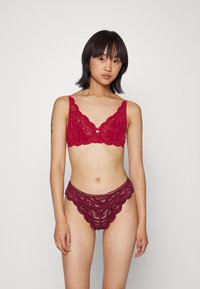 Bralette de renda vermelha com bordas onduladas e um botão central, combinado com panties de renda correspondentes com um padrão intricado e acabamento ondulado.