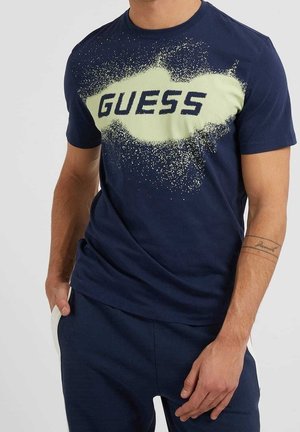 Mężczyzna w granatowej koszulce z jasnym wzorem plam i napisem „GUESS”, w połączeniu z granatowymi spodniami z białymi paskami na kieszeniach.