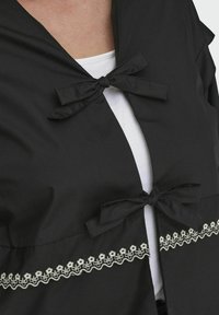 Czarny element odzieży z dwoma materiałowymi kokardkami przy dekolcie oraz dekoracyjnym białym haftem kwiatowym wzdłuż dolnej krawędzi.