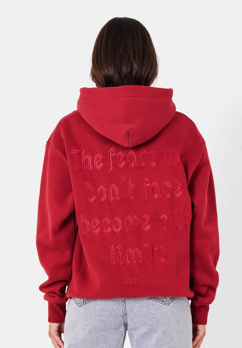 Sweat à capuche rouge avec une poche kangourou, comportant un texte en relief au dos. Manches avec bords côtes et ourlet à bords côtes ; tissu doux avec une coupe décontractée.