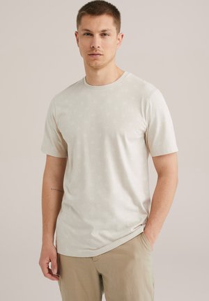 T-shirt a maniche corte, di un leggero beige, con un sottile motivo a diamante, scollo rotondo e tessuto morbido e liscio. Indossato con pantaloni chiari.