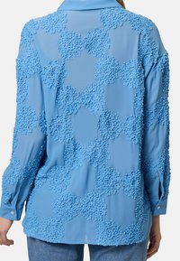 Blouse bleue présentant un design texturé de motifs floraux en relief, manches longues avec des poignets à boutons, et un col de chemise.