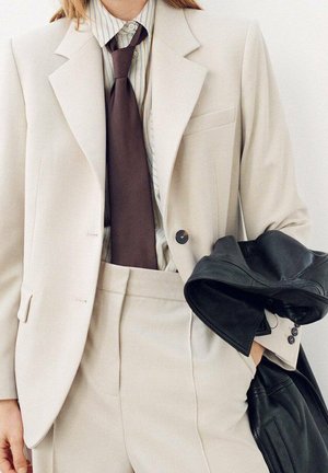Beżowa, dopasowana marynarka z wyciętym klapą, zestawiona z pasiastą koszulą i ciemnobrązowym krawatem. Czarna skórzana kurtka trzymana w jednej ręce.