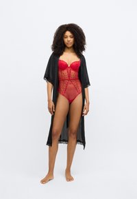 Κόκκινο κορμάκι από δαντέλα με διαφανείς λεπτομέρειες από δίχτυ και μαύρο σατέν κιμονό ρόμπα, με διακόσμηση από δαντέλα. Το μοντέλο στέκεται ξυπόλυτο μπροστά σε λευκό φόντο.