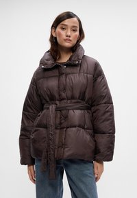 Mlada žena u smešnom tamnosmeđem puffer kaputu s remenom i plavimfarmerkama, koja stoji naslonjena na jednostavnu belu pozadinu.