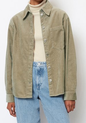 Person trägt ein beigefarbenes Cordhemd mit Knöpfen über einem cremefarbenen, gerippten Rollkragenpullover und hellblaue, hoch taillierte Jeans, die Arme entspannt an den Seiten.