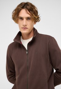 Brown zip-up jacket with a high collar and a soft texture. Features a small logo on the chest and a contrasting inner lining.