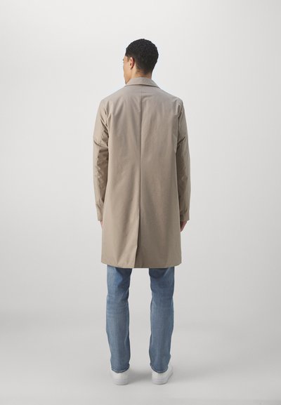 Beige long coat with a collar, back vent, and long sleeves, paired with blue jeans and white sneakers, viewed from the back.