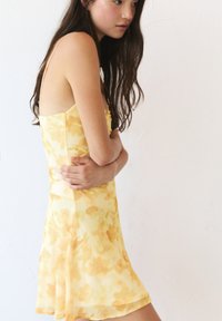 Gelbes Kleid mit Blumenmuster, dünnen Trägern, tailliertem Oberteil und weit ausgestelltem Rock, aus leichtem, durchsichtigem Stoff gefertigt.