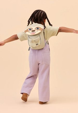 Child with braided hair wearing pastel purple overalls and beige shirt, posing with arms outstretched, carrying a sloth-faced backpack.