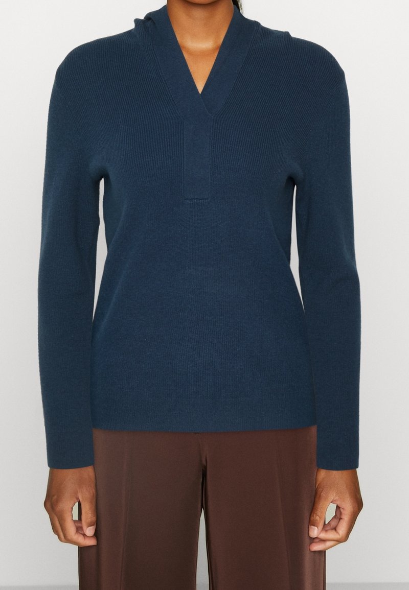 Pull-over en tricot bleu marine avec un col en V et une capuche, présentant une texture côtelée et des manches longues, accompagné d'un pantalon marron.
