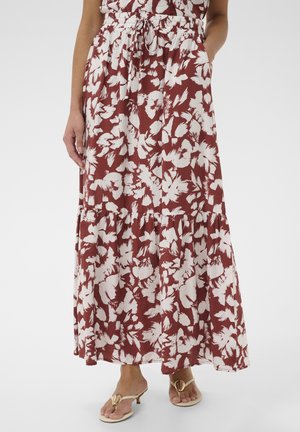 Žena v maxisukni hrdzavej farby s bielym kvetinovým vzorom, elastickým pásom so šnúrkou a béžovými sandálmi na opätku.