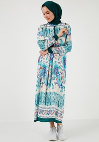 Floral patterned, long-sleeve dress in cream, teal, and pink shades with accent detailing. Paired with white sneakers and a teal hijab.