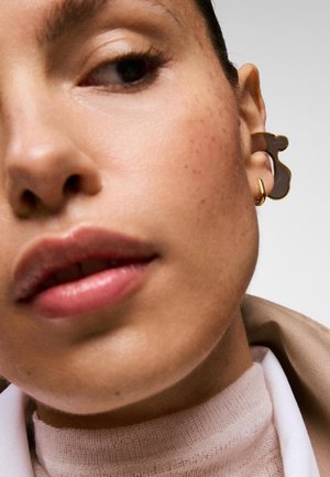 Primer plano de un rostro con pecas claras, que lleva un gran pendiente marrón y dorado y un pequeño aro dorado, vestido con ropa beige y blanca.