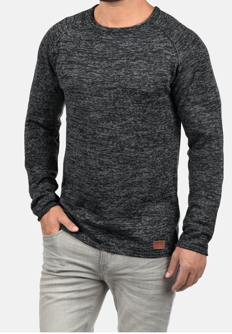 Homme portant un pull en maille texturé à manches longues gris foncé avec un jean gris clair, une main dans la poche, sur fond uni.