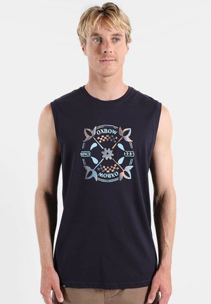 Homme blond portant un débardeur noir avec un motif circulaire et le texte "OXBOW Since 1985", debout devant un fond uni.