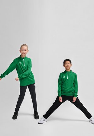 Grüne Sportoberteile mit Reißverschlüssen, kombiniert mit schwarzen Hosen. Beide Kleidungsstücke haben eine glatte Textur und kontrastierende Akzente. Die Schuhe variieren in der Farbe.