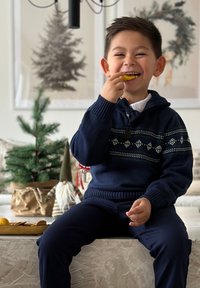 Merihenkinen neulepaita valkoisilla kuoseilla, huppu ja ribbireunukset; hymyilevä lapsi, joka pitää oranssia viipaleita; taustalla joulukoristeita.