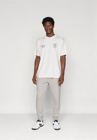 Wit t-shirt met logo's en tekst, grijze joggingbroek en zwart-witte sneakers. Model staat rechtop met een neutrale uitdrukking.